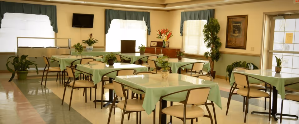 A dining room with several tables covered with light green tablecloths, each table surrounded by beige chairs. The room has large windows with green valances, potted plants on the tables and around the room, a wall-mounted TV, and framed artwork on the walls.