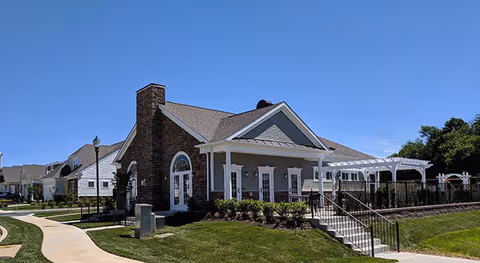 Exterior view of a residential building in the Courts of Clarksburg 55+ Community by Pulte Homes, featuring a brick and siding facade, a covered porch with white columns, a pergola, and a well-maintained lawn under a clear blue sky.
