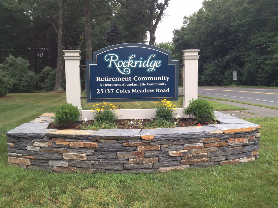 A large blue and white sign for Rockridge Retirement Community, located at 25137 Coles Meadow Road, set on a stone base with small plants and yellow flowers in front. The sign is positioned near a road with trees and greenery in the background.
