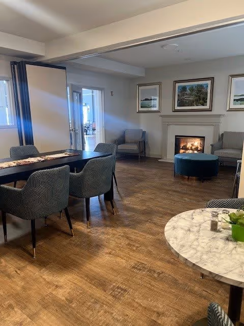 A cozy common area with wooden flooring featuring a long table with six patterned chairs, a round marble table with a small plant and salt and pepper shakers, two armchairs near a lit fireplace, and framed artwork on the walls.
