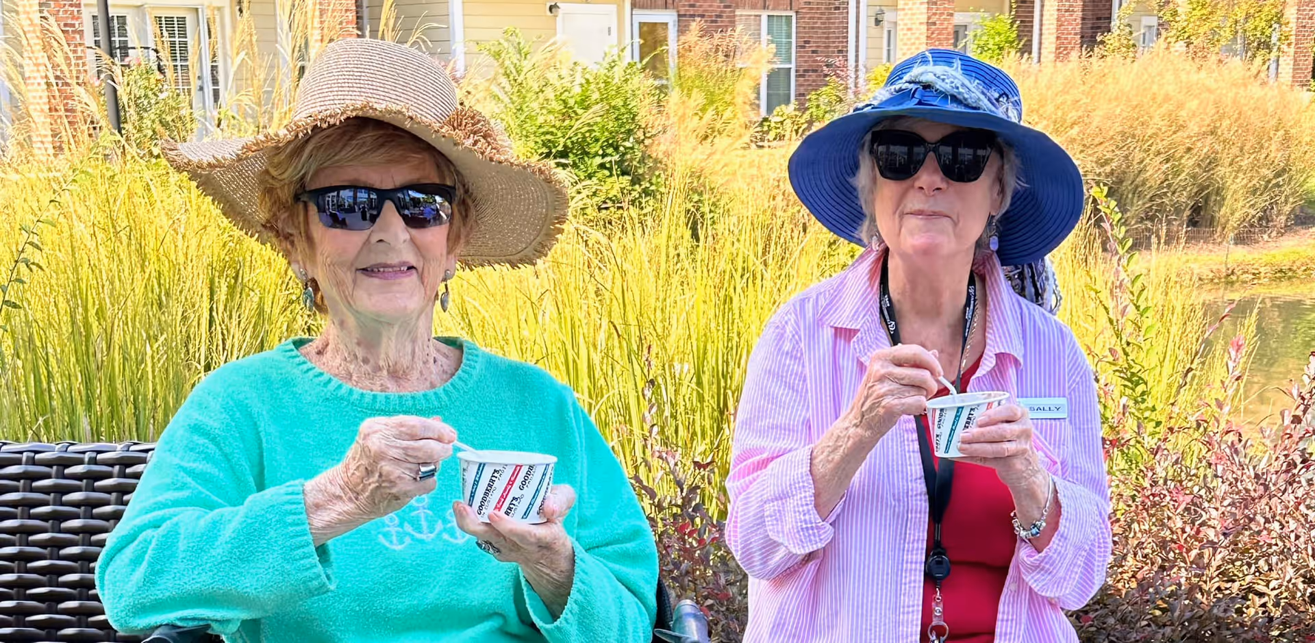 Two elderly women sitting outdoors enjoying ice cream. Both are wearing sunglasses and wide-brimmed hats. Behind them are tall grasses and a pond, with brick buildings in the background.