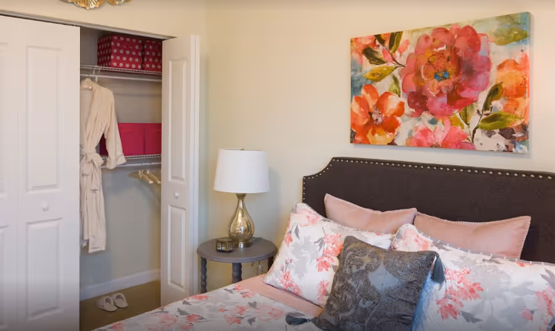 A decorated bedroom with a floral bed and pillows, a nightstand with a lamp, colorful floral wall art, and an open closet showing robes and storage boxes.