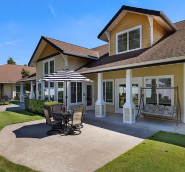 Outdoor patio area of a senior living facility with a table and chairs under a striped umbrella, a cushioned swing, and a yellow building with white trim and multiple windows in the background.