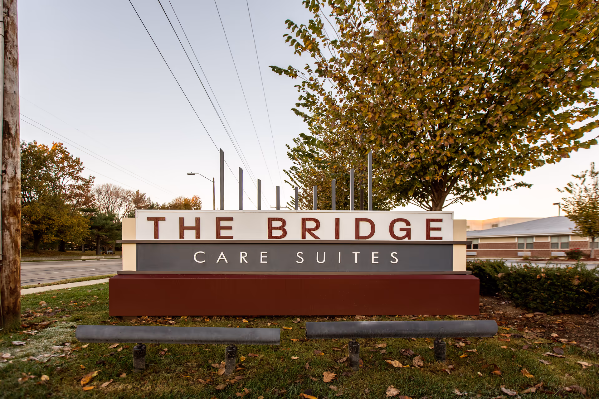 Outdoor sign for The Bridge Care Suites facility with trees and a street in the background during daytime.
