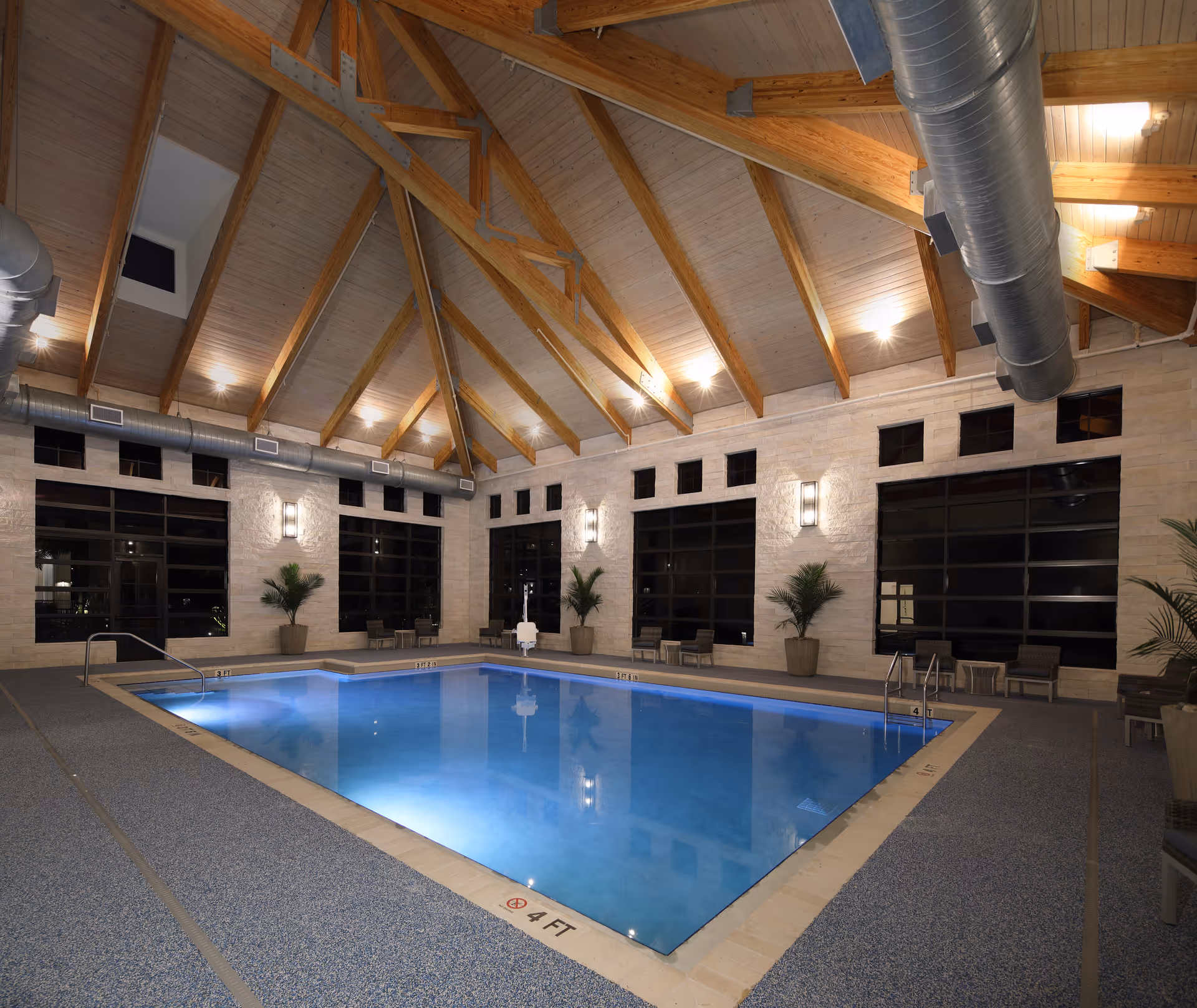 Indoor swimming pool with clear blue water in a room with high wooden vaulted ceiling and exposed beams. The walls are light-colored stone with large black-framed windows. Several potted plants and chairs are arranged around the pool area. The pool depth is marked as 4 feet.