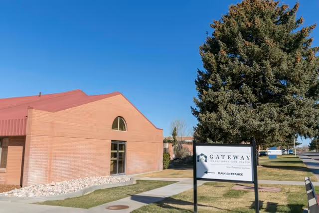 Exterior view of Gateway Transitional Care Center building with a brick facade and a red roof under a clear blue sky. A sign in the foreground indicates the main entrance with an arrow pointing left. There is a large tree to the right of the sign and a sidewalk leading to the entrance.