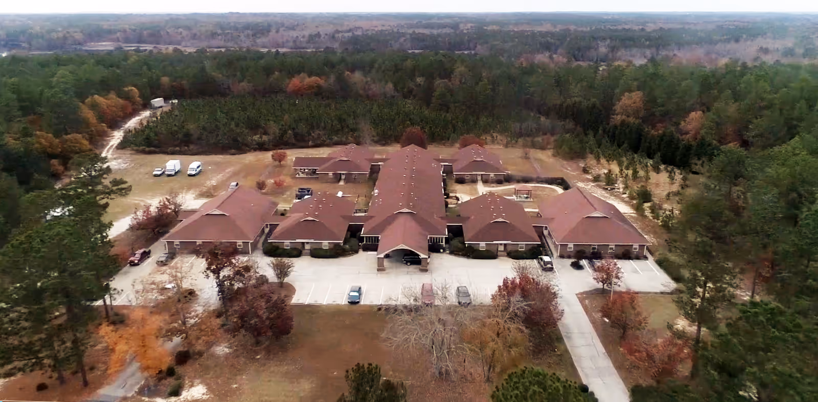Aerial view of Pinedale Residential Center, a large single-story building complex with multiple wings and a red roof, surrounded by trees and greenery. There is a parking lot with several vehicles in front of the building and a driveway leading to the entrance.