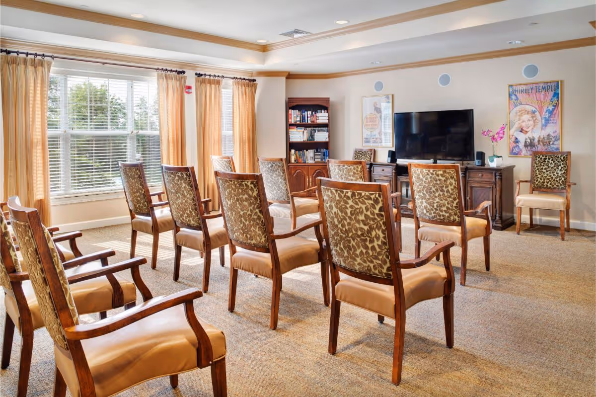 Sunlit communal activity room with rows of patterned wooden chairs facing a TV, bookshelf and large windows with curtains.