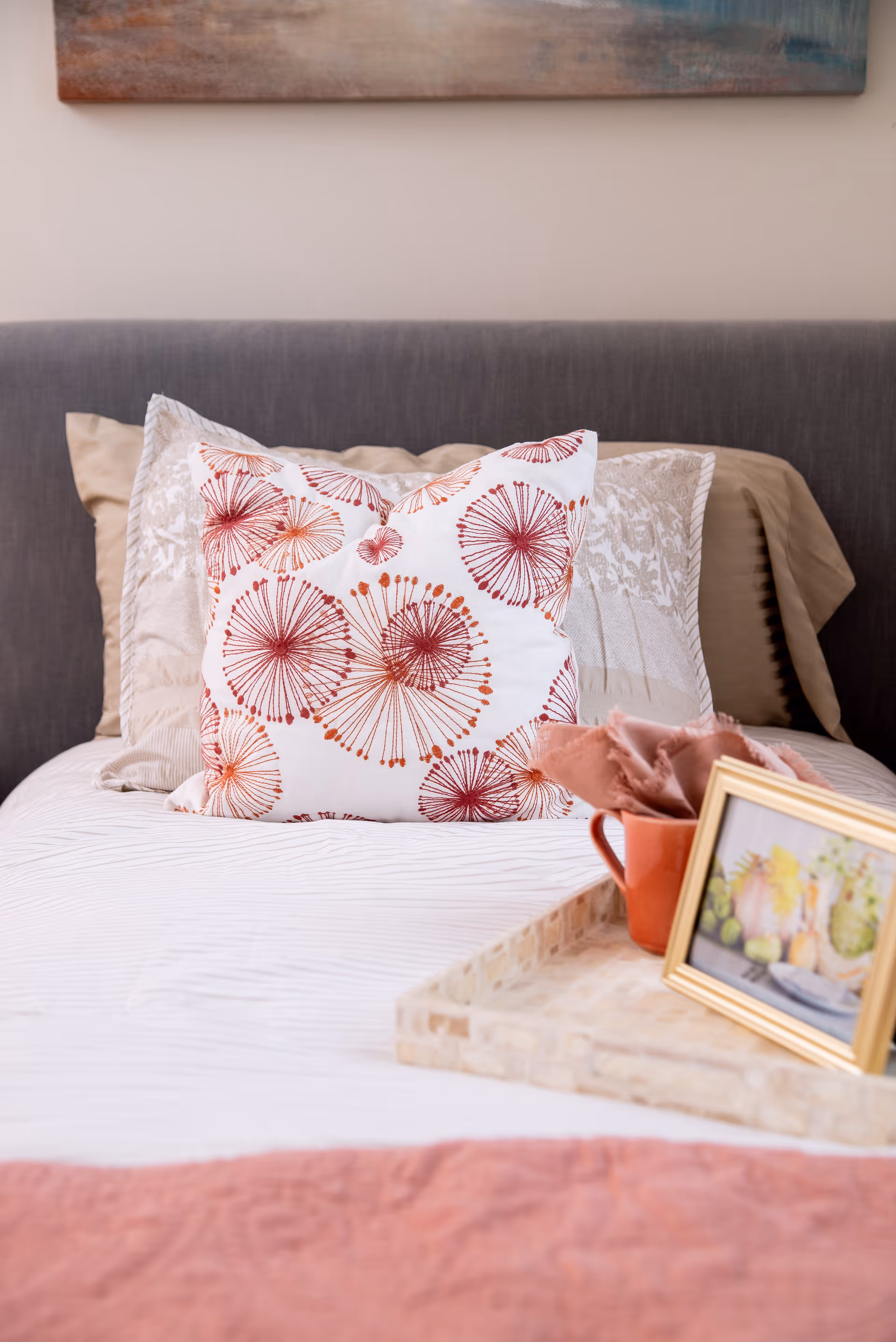 Close-up of a neatly made bed with a grey upholstered headboard, featuring decorative pillows including one with a red and orange floral pattern. A beige and white patterned pillow is behind it. On the bed is a tray holding a red mug with a cloth napkin and a small framed picture with a floral design. A piece of artwork is partially visible on the wall above the headboard.