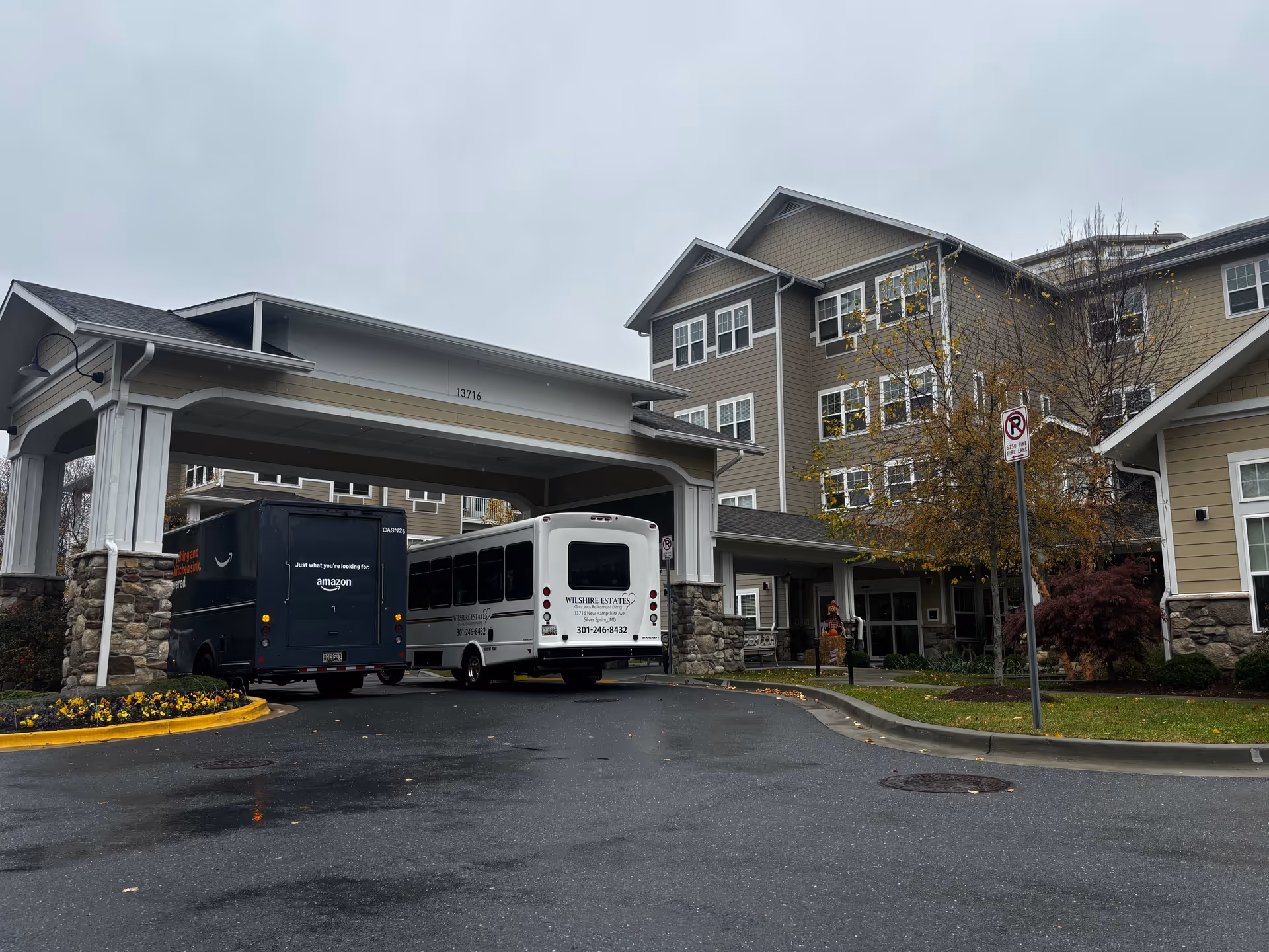 Exterior view of Wilshire Estates Gracious Retirement Living building on a cloudy day with a covered entrance area. Two vehicles, an Amazon delivery truck and a Wilshire Estates shuttle bus, are parked under the covered entrance. There are trees with autumn leaves and a no parking fire lane sign near the entrance.