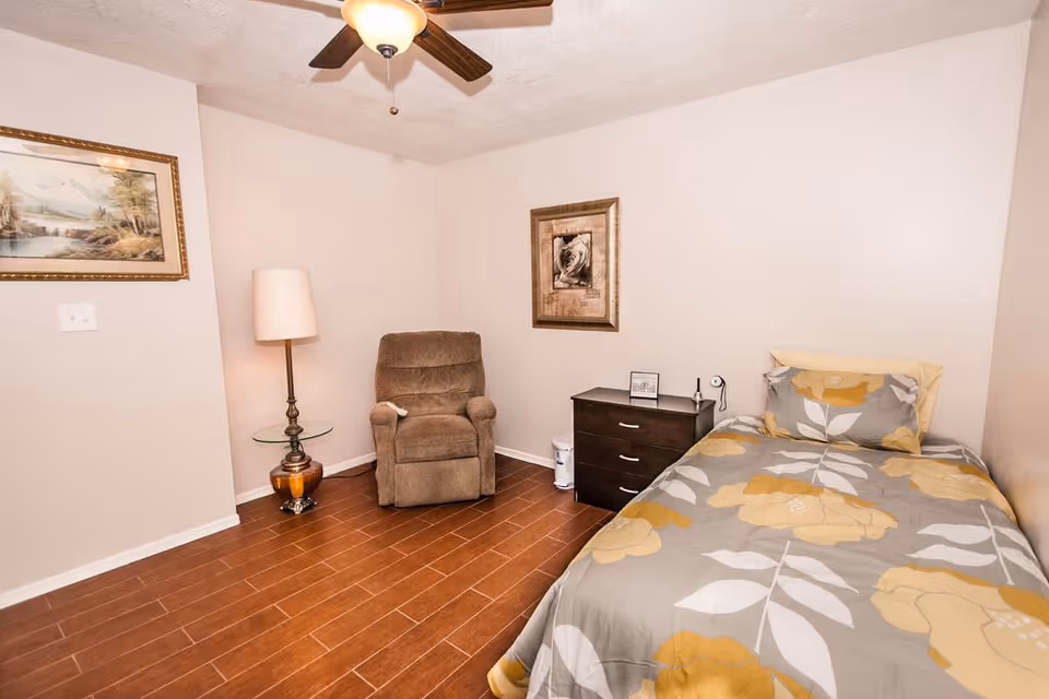 A small bedroom with a single bed covered in a gray and yellow floral bedspread, a dark wooden nightstand with a small lamp and framed picture, a brown recliner chair, a floor lamp with a glass side table, two framed pictures on the walls, and a ceiling fan with a light fixture. The floor is wooden and the walls are painted light beige.