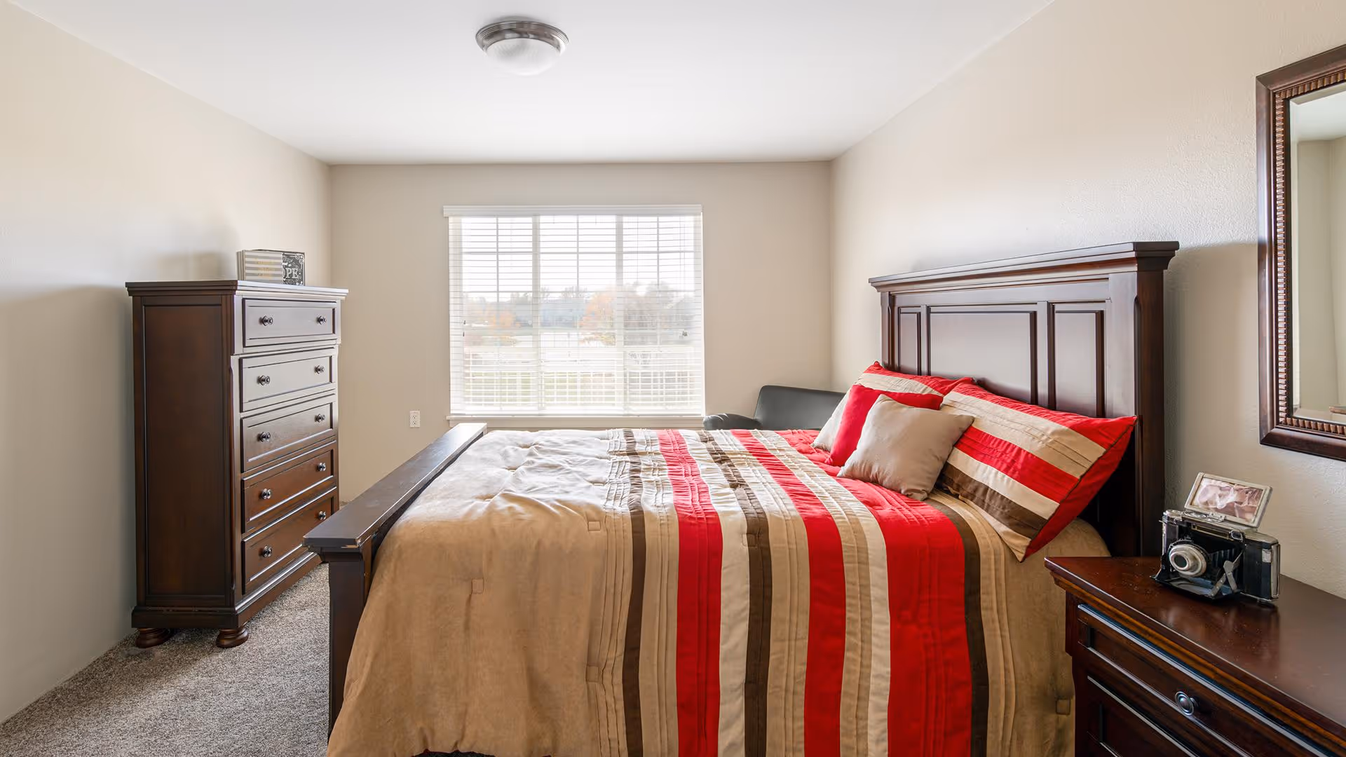 A bedroom with a large wooden bed featuring a red, beige, and brown striped comforter and matching pillows. There is a wooden dresser with drawers on the left side and a wooden nightstand with a vintage camera and a framed photo on the right side. A large window with blinds is centered on the back wall, letting in natural light. A mirror hangs on the wall above the nightstand.