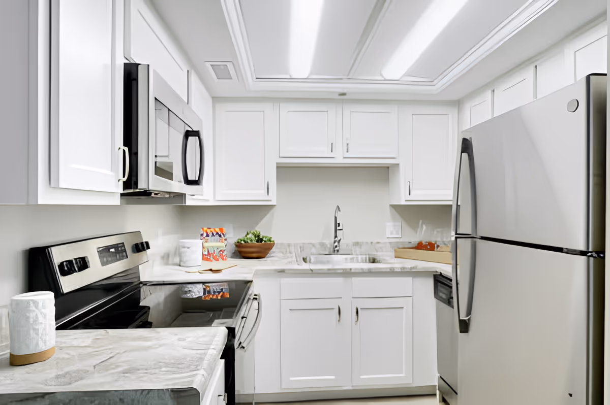 A modern kitchen with white cabinets, a marble countertop, a stainless steel refrigerator, microwave, stove, and a sink. There is a bowl with green leafy vegetables and some packaged items on the counter.