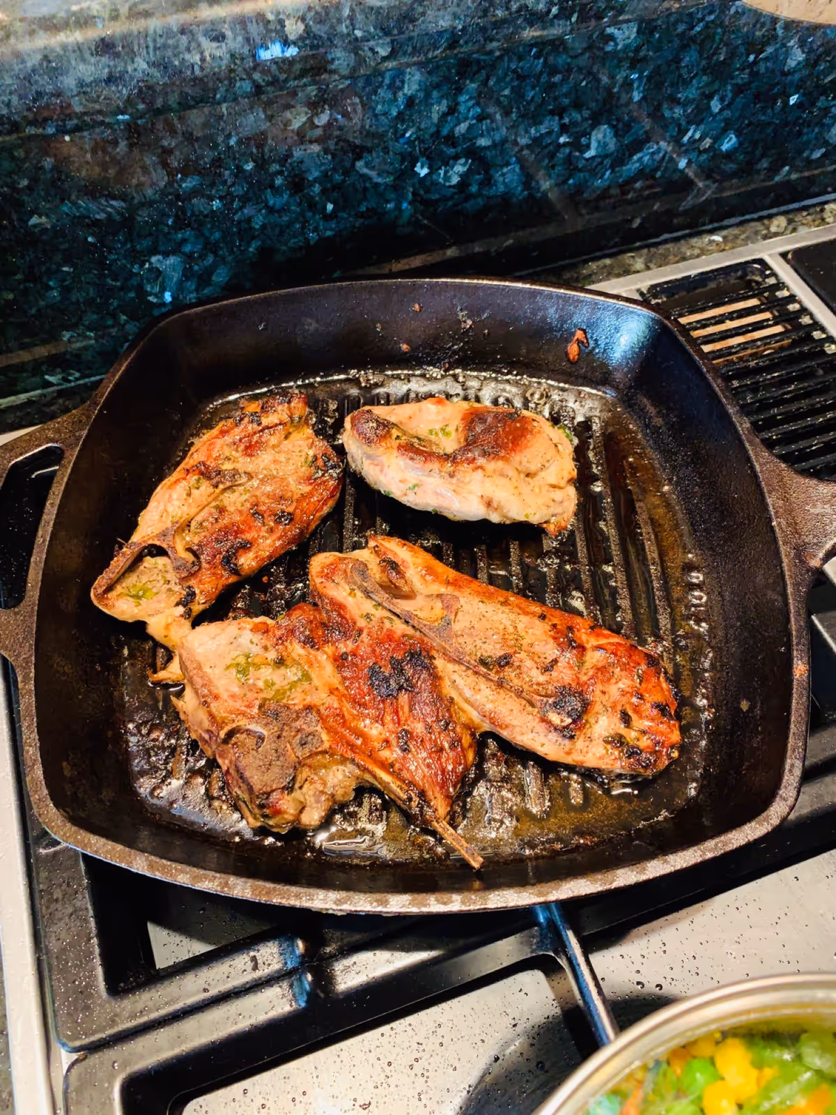 Four pieces of seasoned meat cooking on a black cast iron grill pan on a stovetop with a granite backsplash. A pot with mixed vegetables is partially visible in the bottom right corner.