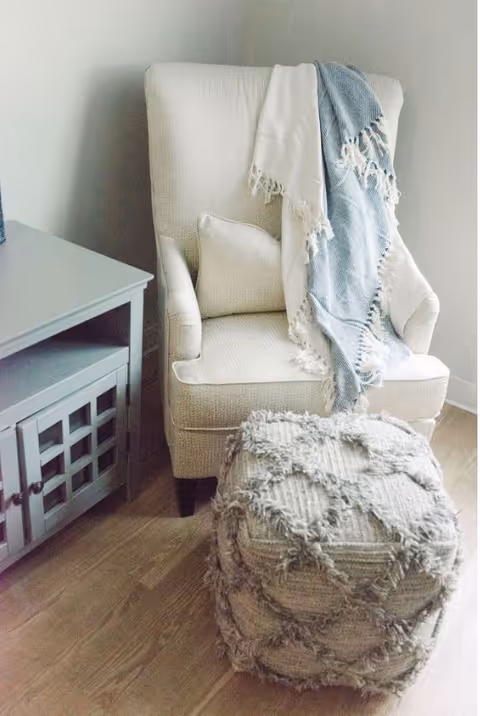A cozy beige armchair with a matching cushion and a blue and white fringed throw draped over the back. In front of the chair is a textured pouf with a diamond pattern. To the left of the chair is a gray wooden cabinet with glass-paneled doors. The floor is light wood.