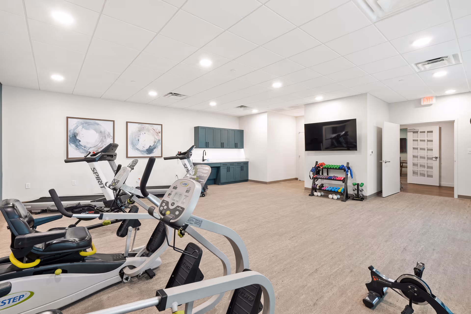 A bright and spacious fitness room with exercise equipment including stationary bikes and a rowing machine. The room features light-colored walls, a wood-like floor, two abstract paintings on the wall, a wall-mounted TV, and a rack with various dumbbells. There is a kitchenette area with green cabinets and a sink, and multiple doors leading to other rooms.