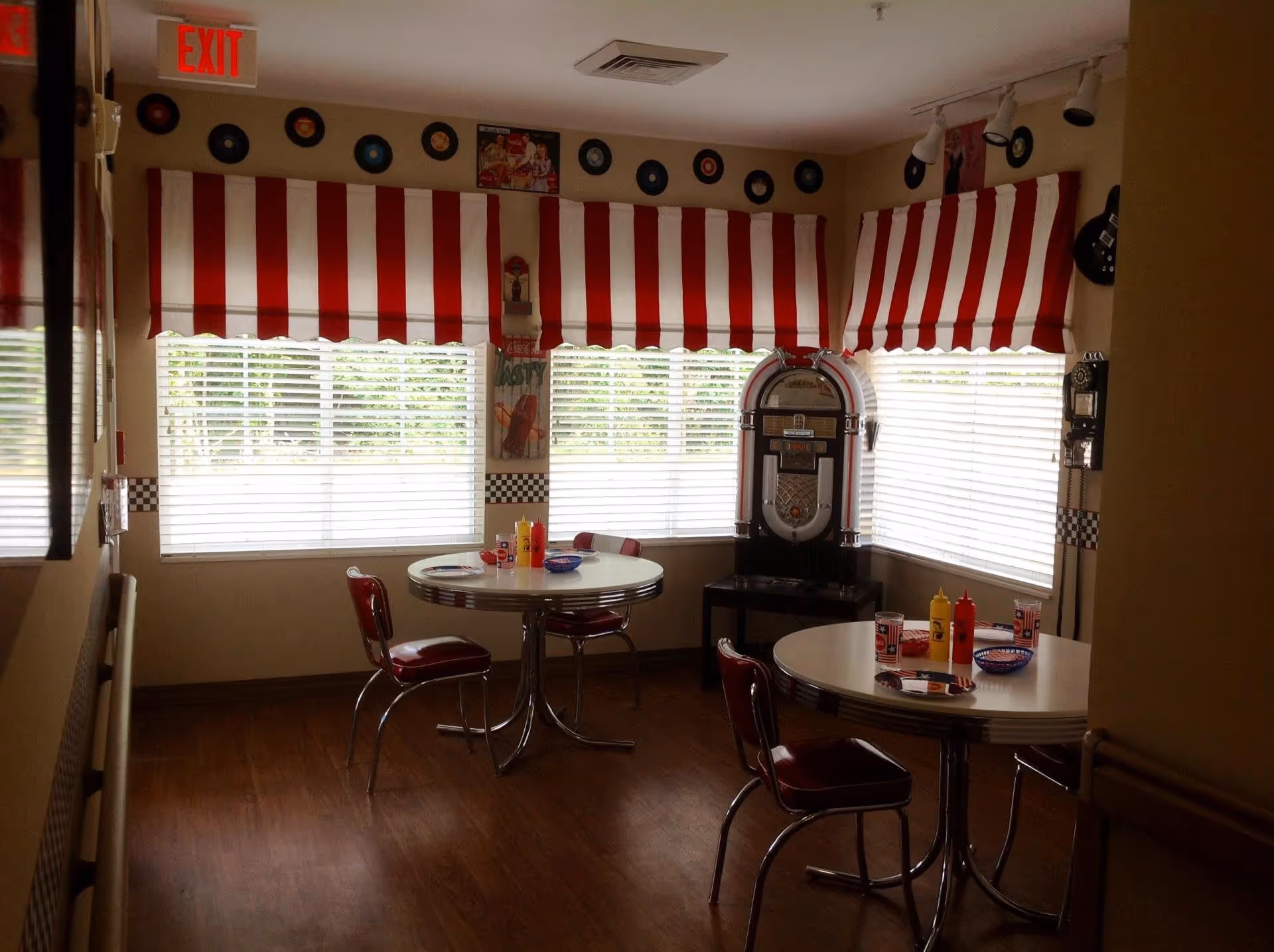 A retro-style dining area with two round white tables and red cushioned chairs. The windows have red and white striped valances, and the walls are decorated with vinyl records and vintage posters. A jukebox and a vintage wall phone are visible in the corner. The floor is wooden, and there are condiment bottles on the tables.