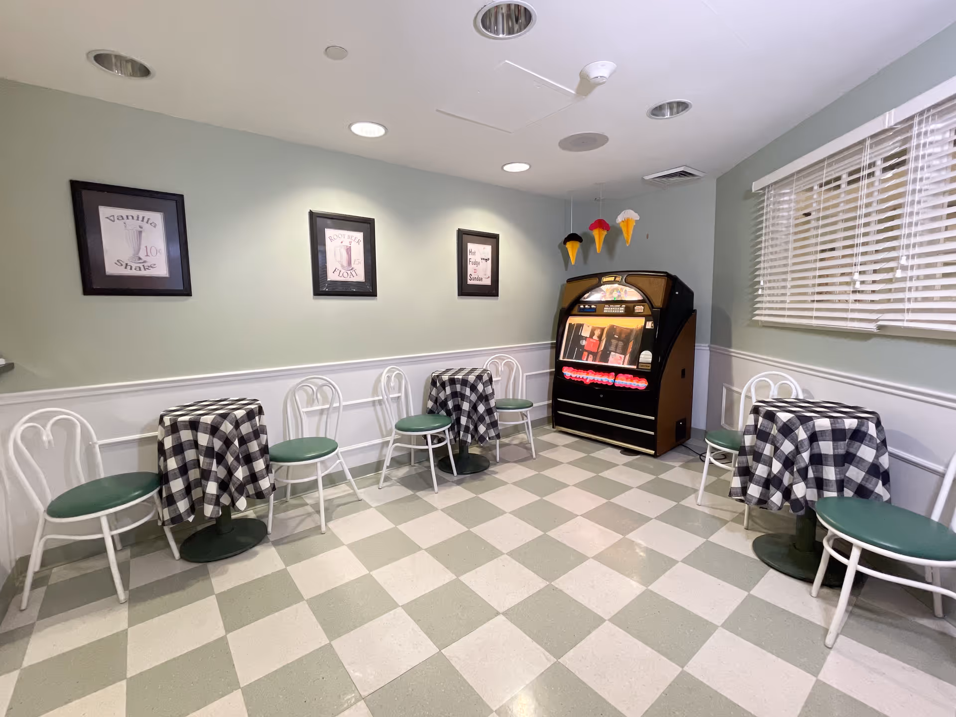 A small dining area with three tables covered in black and white checkered tablecloths, each surrounded by white chairs with green seats. The room has light green walls with white wainscoting and three framed vintage-style posters on the wall. There is a jukebox in the corner with three large decorative ice cream cones hanging above it. A window with white blinds is on the right side of the room.