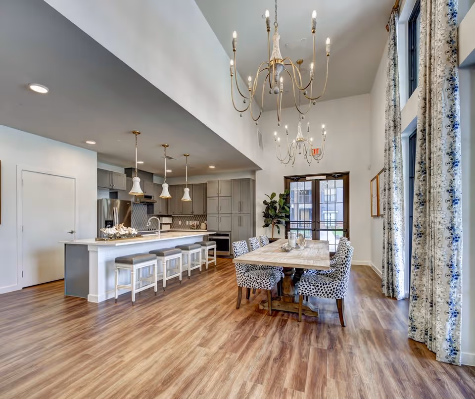 A spacious, modern kitchen and dining area with wood flooring. The kitchen features gray cabinets, stainless steel appliances, a large island with a sink, and four bar stools. The dining area has a wooden table surrounded by six patterned chairs, with tall windows covered by patterned curtains. Two elegant chandeliers hang from the high ceiling, and a glass door leads outside.