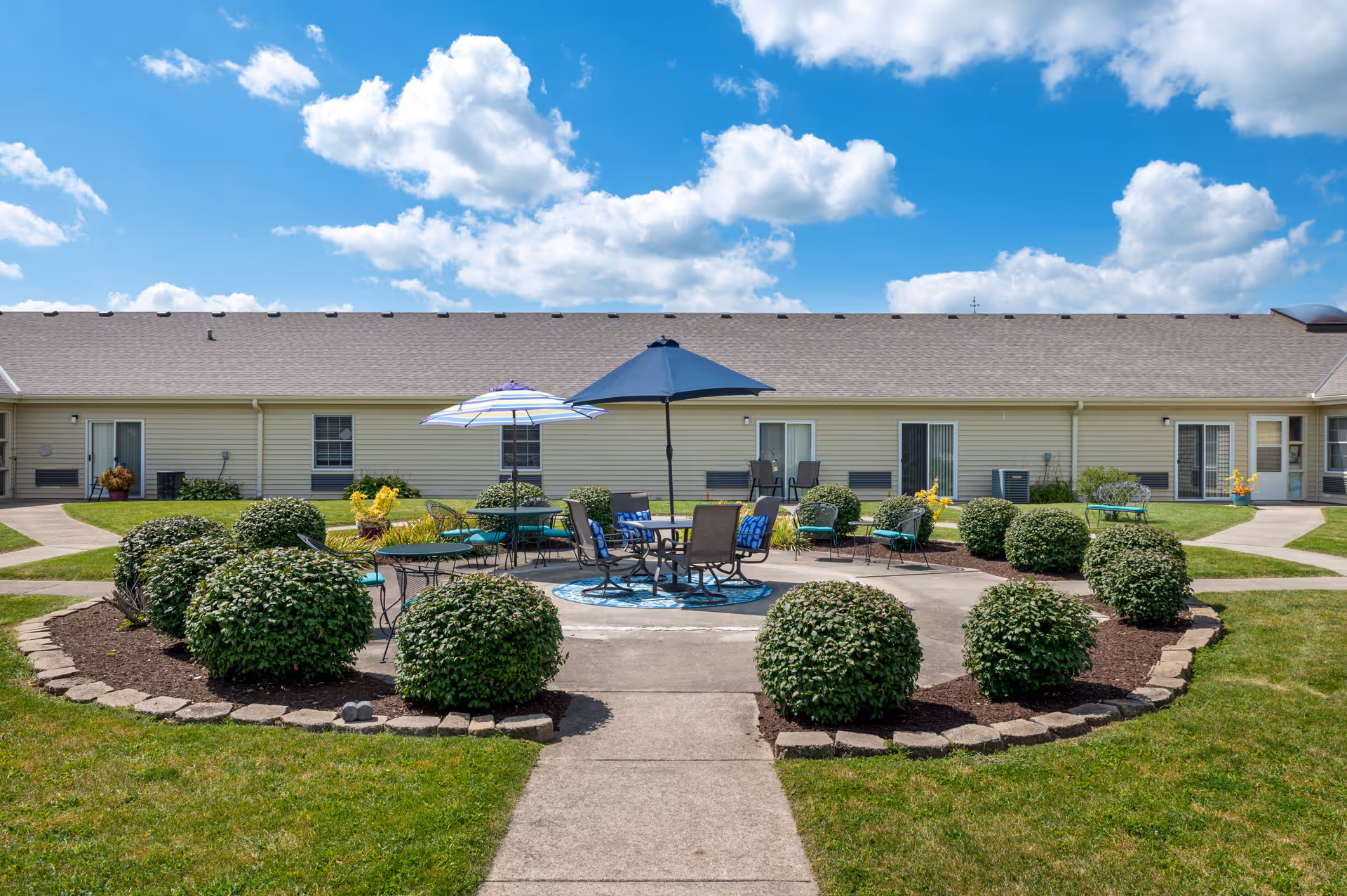 Outdoor seating area in a garden courtyard of Oakley Courts Assisted Living Community with round tables, chairs, and umbrellas surrounded by neatly trimmed bushes and pathways, under a blue sky with scattered clouds.