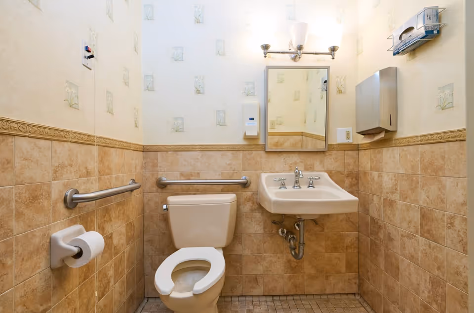 A small bathroom with beige tiled walls and floor, featuring a toilet with grab bars on both sides, a wall-mounted sink with exposed plumbing, a mirror above the sink, a soap dispenser, a paper towel dispenser, and a toilet paper holder.