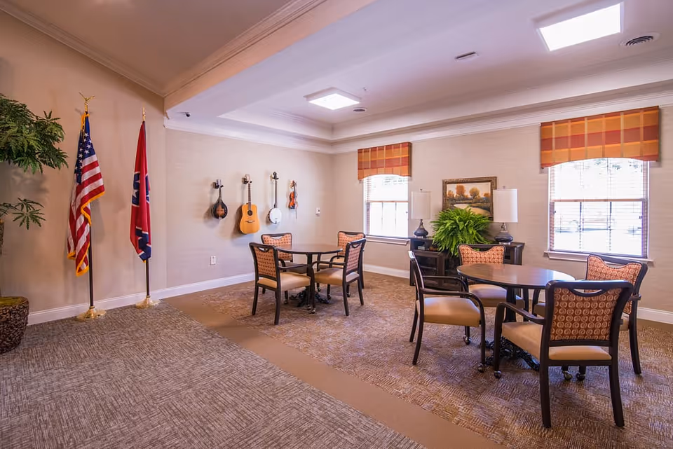 A well-lit common area with two round tables, each surrounded by four cushioned chairs. On the wall, there are four stringed instruments hanging, including a guitar and a banjo. Two windows with orange and yellow plaid valances let in natural light. There are two flags standing in the corner, an American flag and a Tennessee state flag. A large potted plant is near the flags, and a sideboard with two lamps and a green plant is positioned between the windows. The room has a carpeted floor and a neutral color scheme.