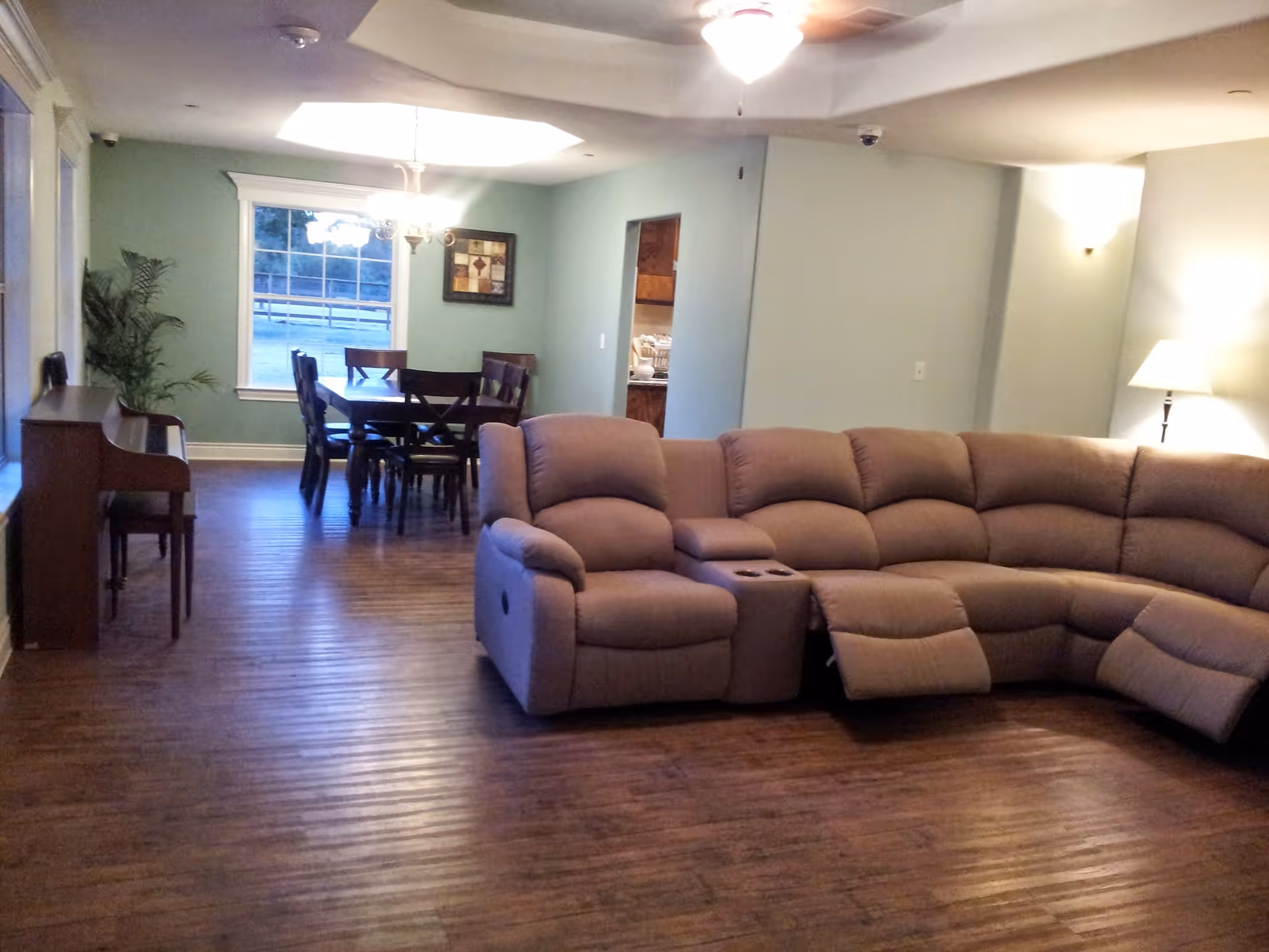 A spacious living area featuring a large beige sectional recliner sofa with cup holders, a wooden floor, a piano with a bench along the left wall, and a dining area with a wooden table and six chairs near a window. The walls are painted light green, and the room is lit by ceiling lights and a floor lamp.