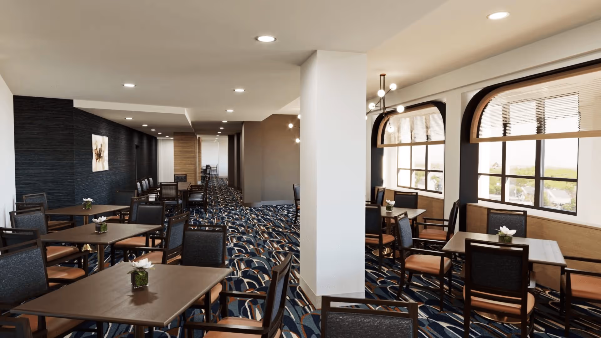 Interior view of a dining room with multiple tables and chairs arranged neatly. The room has large windows with arched frames and blinds, allowing natural light to brighten the space. The floor is covered with a patterned carpet in shades of blue, brown, and beige. Modern light fixtures hang from the ceiling, and small flower arrangements are placed on each table.