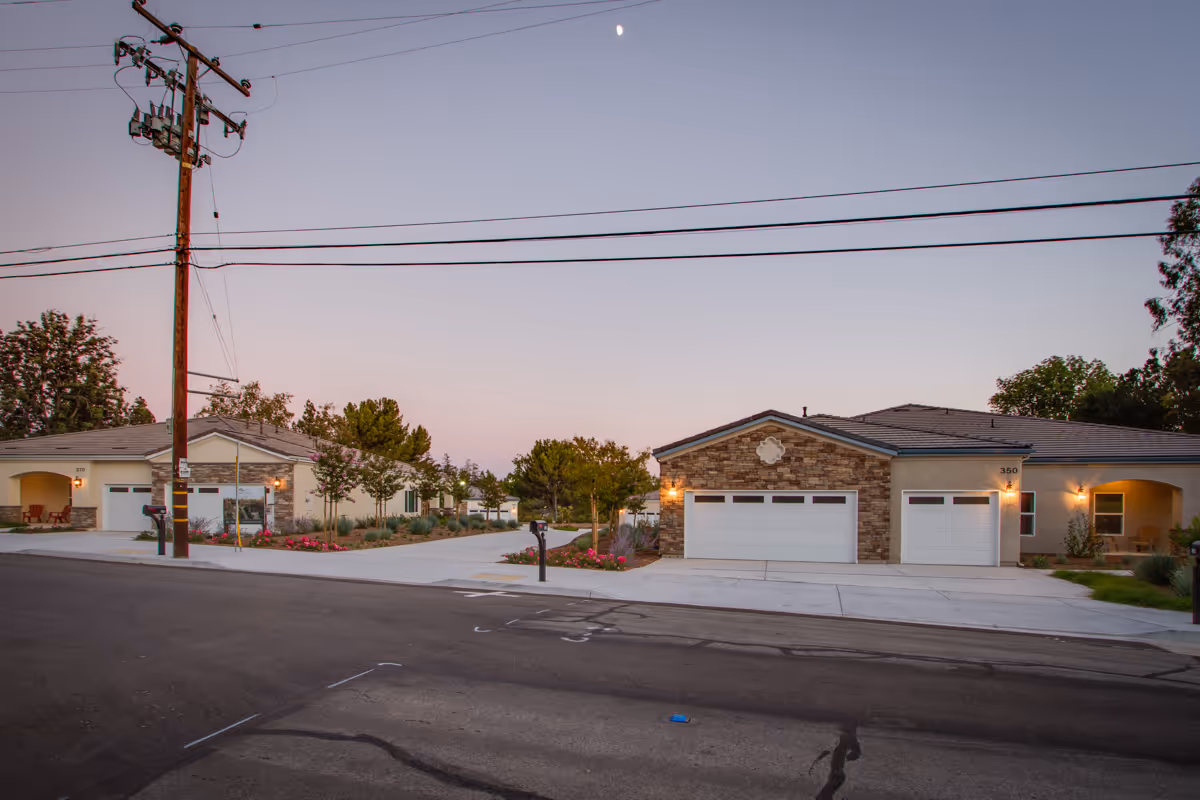 Exterior view of a senior living facility at dusk showing two single-story buildings with garages, well-maintained landscaping, and a clear sky with a visible crescent moon.