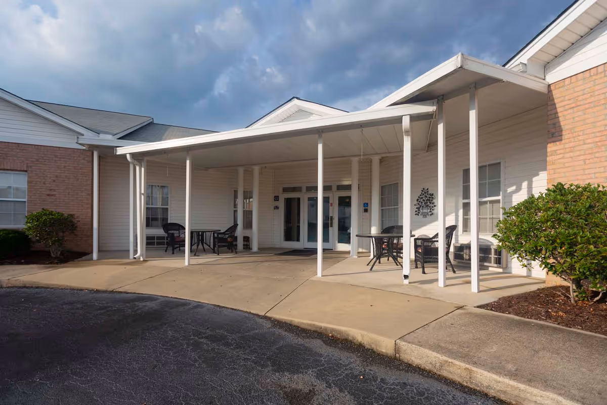 Covered entrance area of a building with white pillars, outdoor seating including tables and chairs, and a brick and white siding exterior under a cloudy sky.