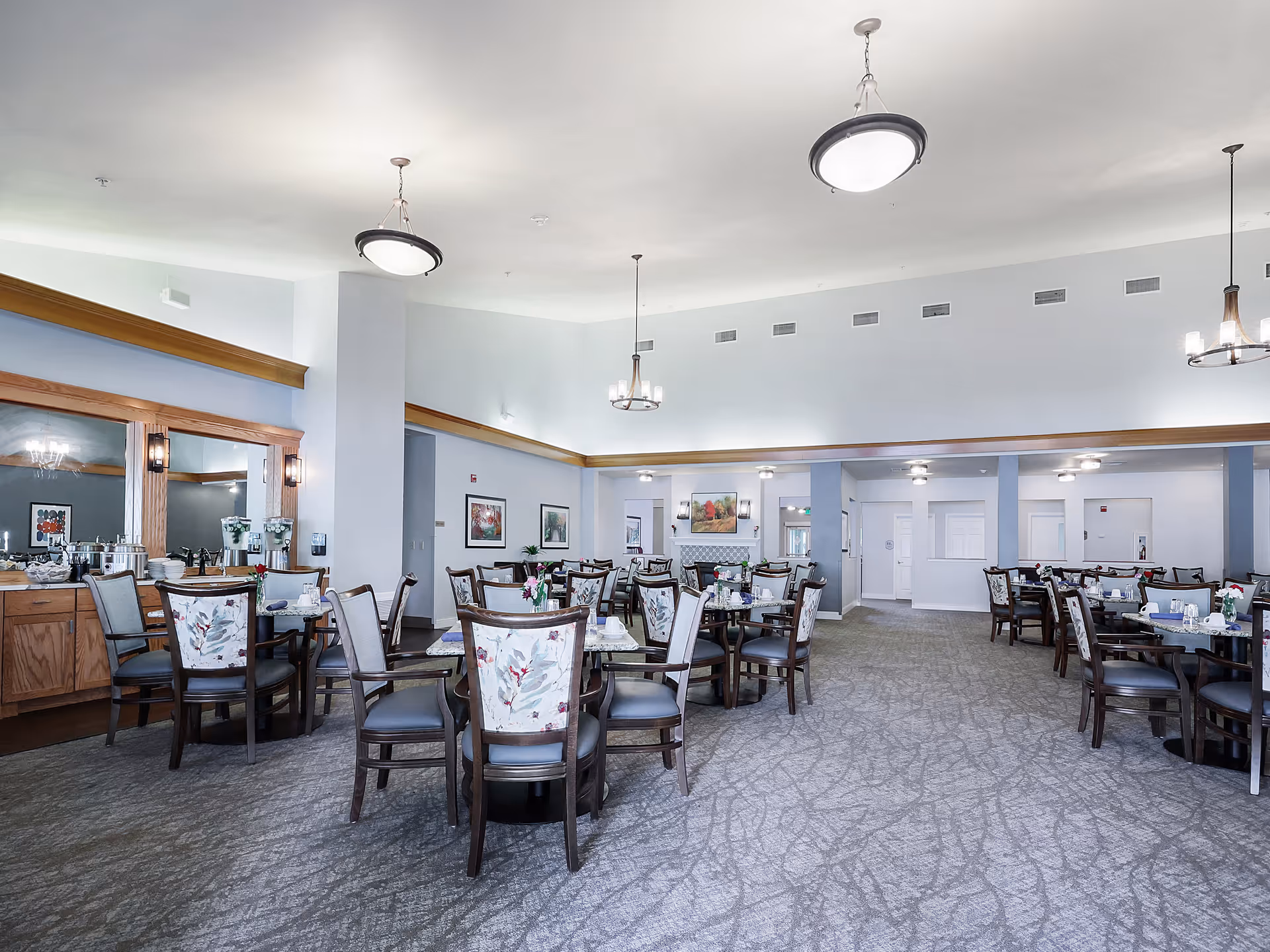 A spacious dining room in a senior living facility with multiple round tables and chairs arranged neatly. The chairs have floral patterned backs and dark wooden frames. The room features soft lighting with several ceiling light fixtures and wall sconces. There is a large mirror on one wall above a wooden buffet area with water dispensers and other dining essentials. The carpet has a subtle pattern, and framed artwork decorates the walls.
