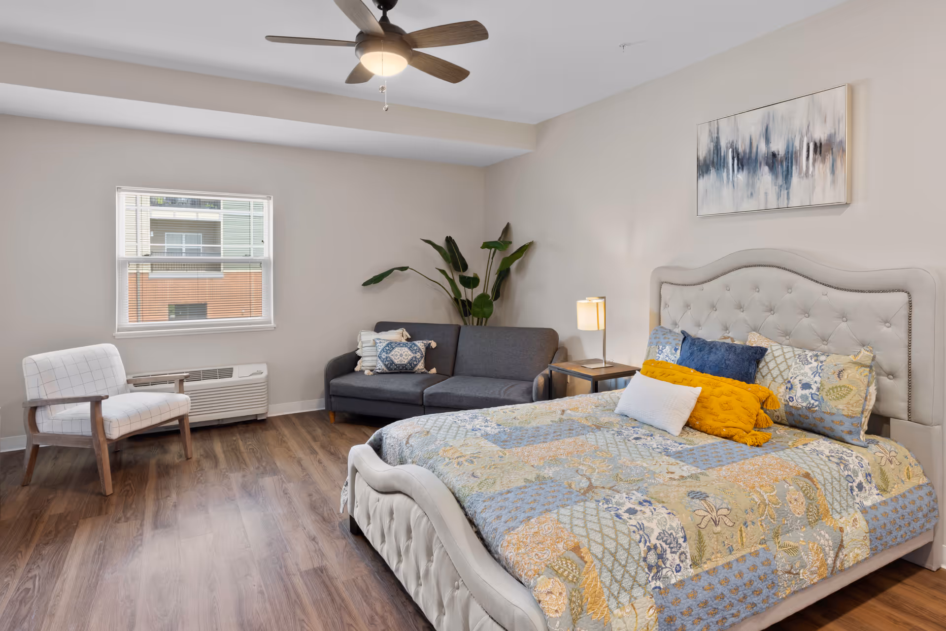 A bright and cozy bedroom featuring a large upholstered bed with a colorful quilt and multiple pillows. To the right of the bed is a small side table with a lamp. Against the far wall is a dark gray loveseat with decorative pillows and a tall green plant behind it. A white armchair with wooden arms is positioned near a window that lets in natural light. The room has wood flooring, a ceiling fan with a light, and a modern abstract painting above the bed.