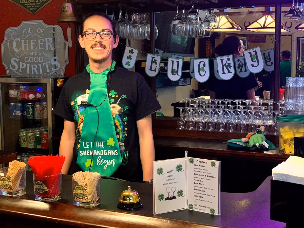 A smiling man wearing glasses and a green tie with shamrocks stands behind a bar counter decorated with a 'LUCKY' banner. The bar has glasses hanging overhead, a small menu with wine and cocktail options, and containers with red and beige straws. Behind the man, there is a sign that reads 'Full of Cheer and Good Spirits' and a refrigerator stocked with beverages.