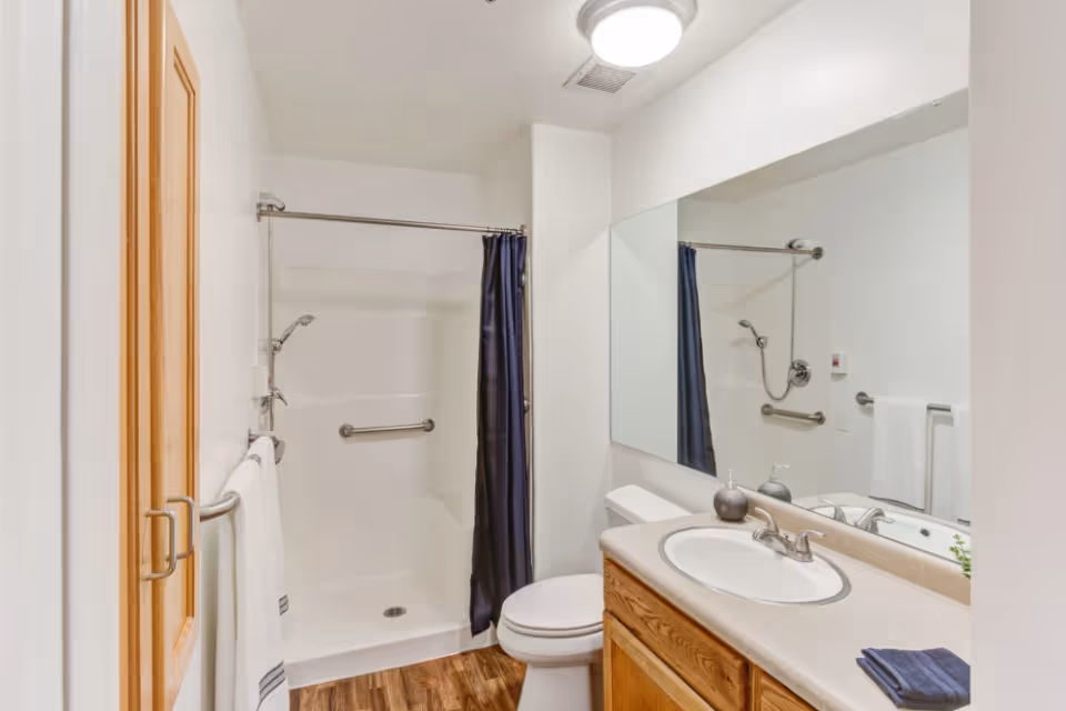 A clean bathroom with a shower area featuring a silver showerhead and a dark blue shower curtain. There is a white toilet next to a wooden vanity with a white sink and a large mirror above it. The bathroom has white walls and a wooden floor, with a towel hanging on a silver towel rack.