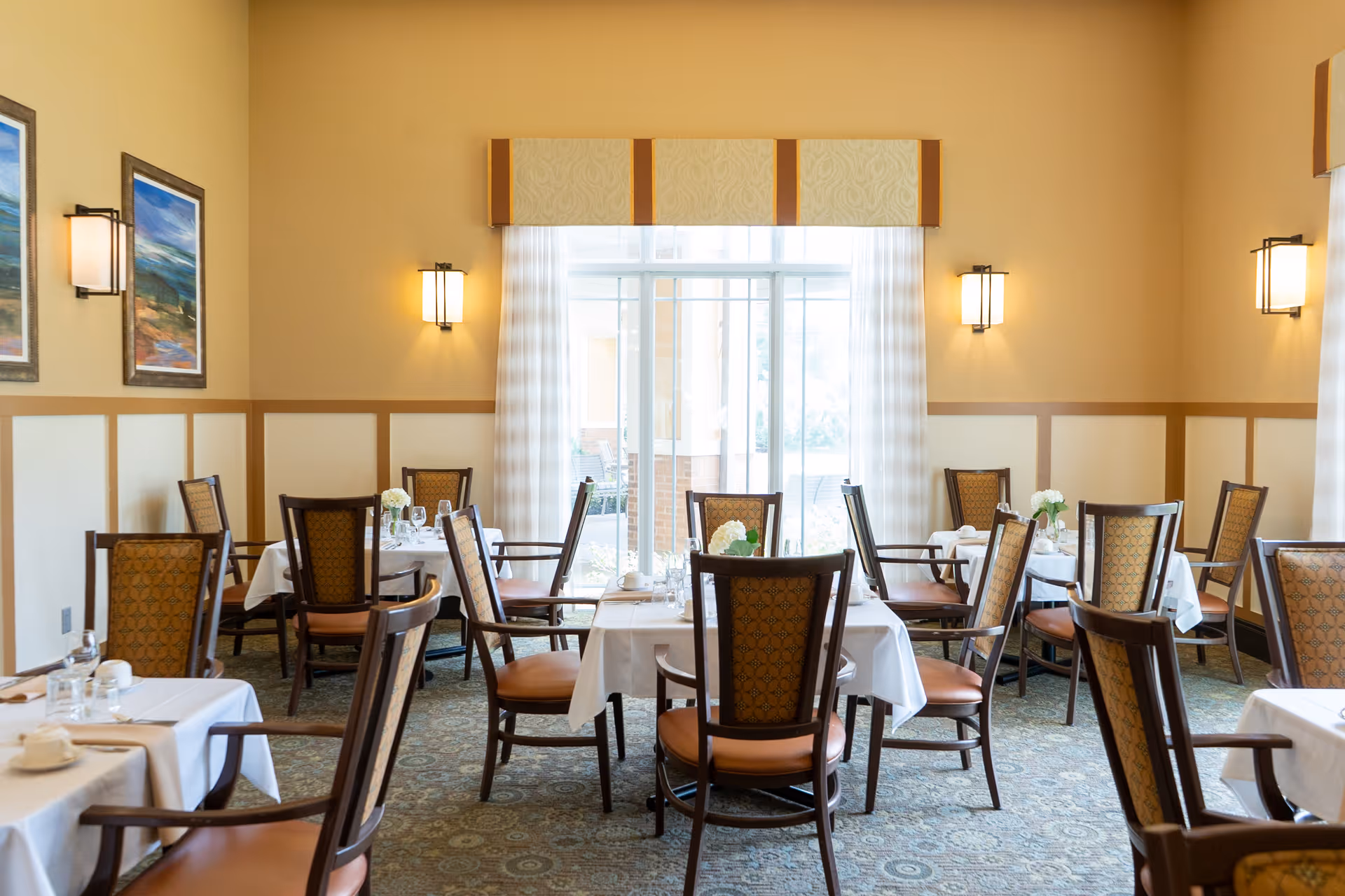 Dining room with multiple set tables and wooden chairs arranged around a large central window.
