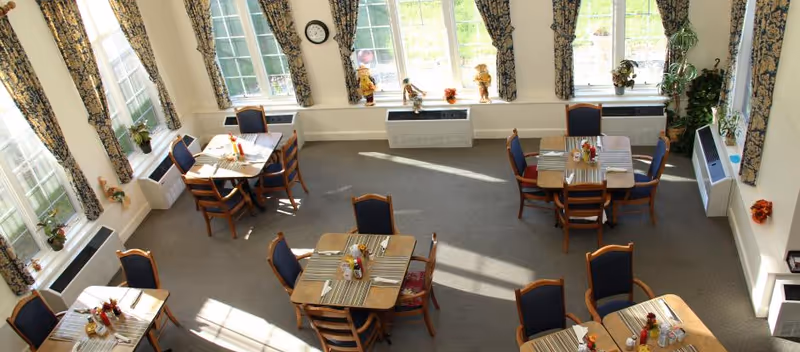 Sunlit communal dining room with several set wooden tables and chairs beside large windows with floral curtains.