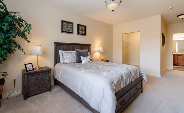 A neatly made bed with a wooden headboard and floral bedding in a bedroom. There are two bedside tables with lamps on each side of the bed. A framed photo is on the left bedside table. The room has beige walls and carpeted floor. A large plant is in the corner to the left. An open doorway leads to a walk-in closet and another doorway leads to a bathroom with a vanity and mirror visible.