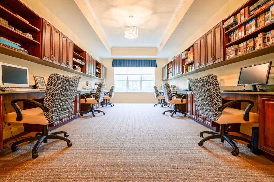 A bright, spacious computer room with a row of desks and patterned swivel chairs on both sides. Each desk has a computer monitor, and above the desks are wooden cabinets and shelves filled with books and board games. A large window at the end of the room lets in natural light, and a ceiling light fixture is centered above.