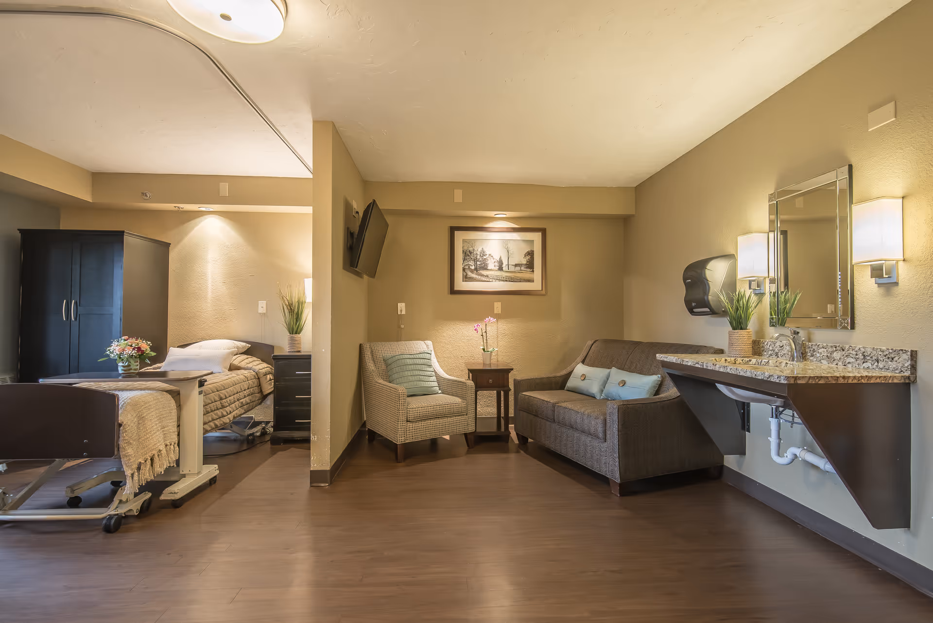 A cozy senior living room and bedroom area with a hospital-style bed, a dark wooden wardrobe, a small table with flowers, a comfortable armchair, a sofa with cushions, a wall-mounted TV, a framed picture on the wall, and a granite countertop with a sink and mirror. The room has warm lighting and wooden flooring.