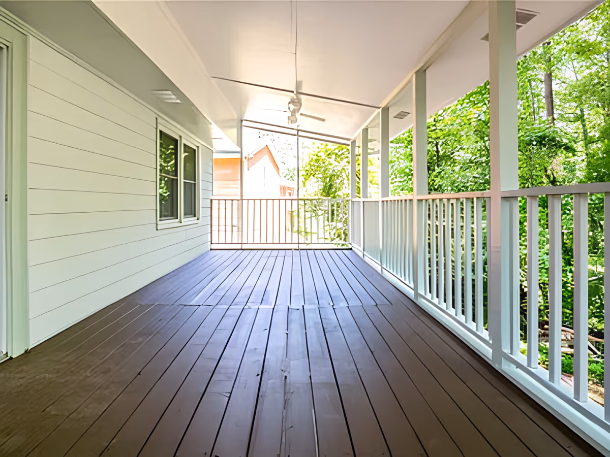 A spacious covered porch with brown wooden flooring and white railings. The porch has a white ceiling with a ceiling fan and overlooks green trees and a neighboring house.