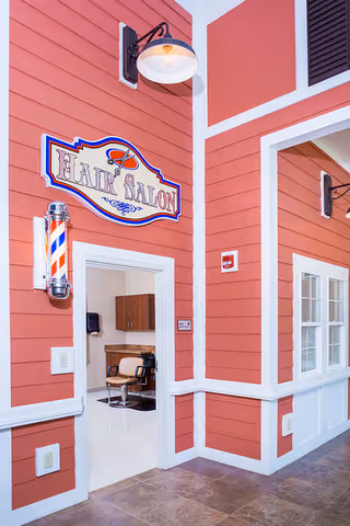 Interior view of a hair salon area within a facility, featuring a red and white exterior-style wall with a vintage barber pole and a sign that reads 'Hair Salon'. Inside, a salon chair and cabinetry are visible.