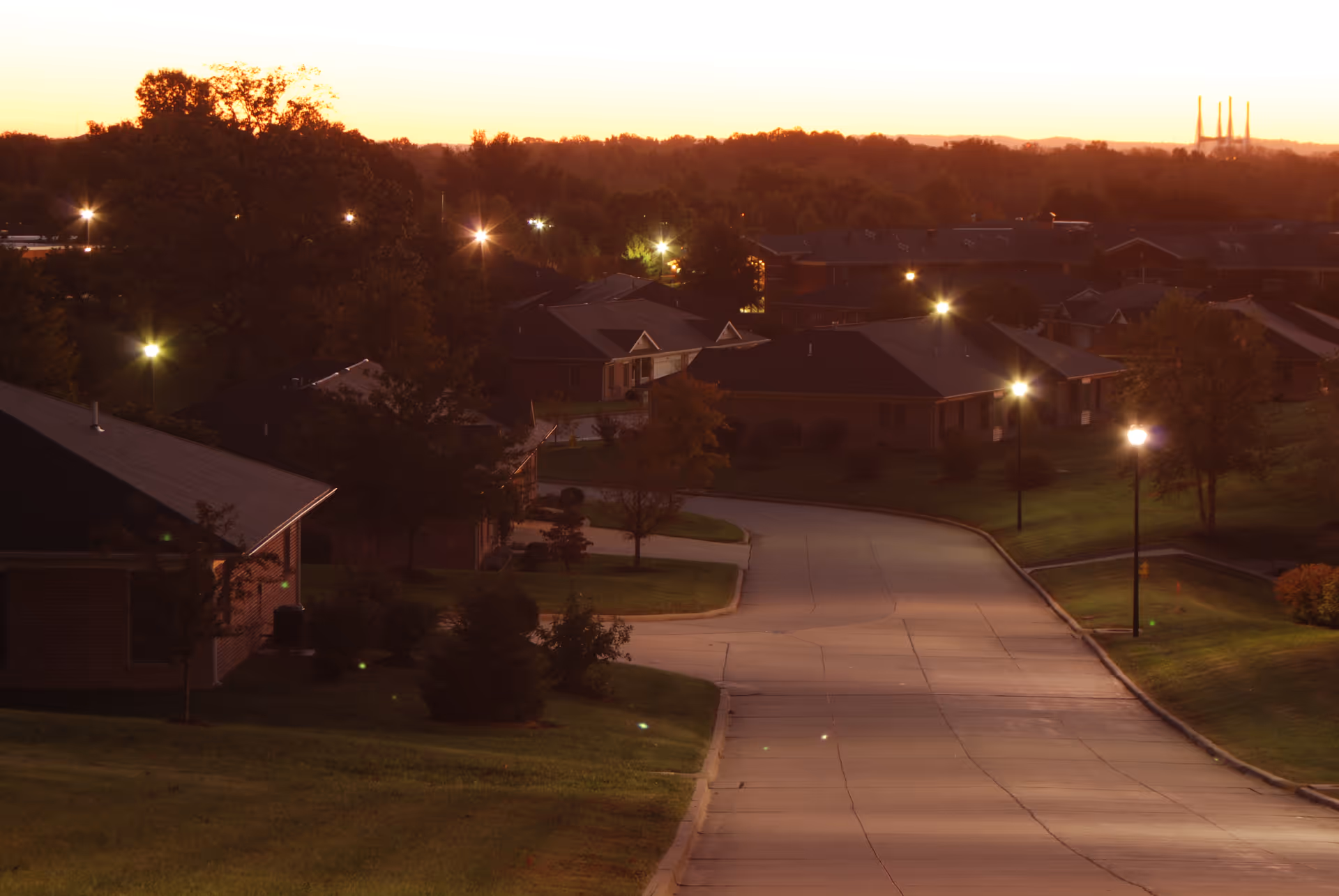 Dusk view of a quiet curving road lined with single-story retirement community buildings and lit streetlights.