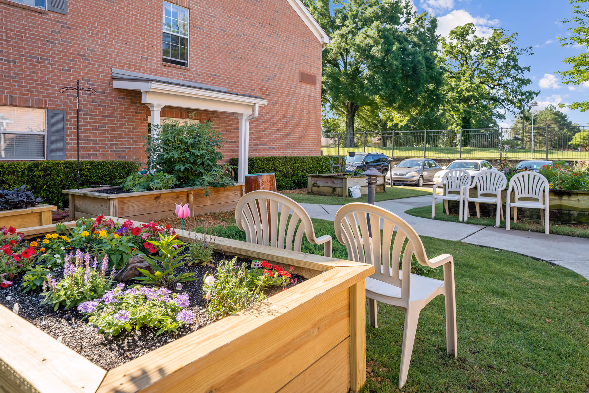 Outdoor garden area at Town Village Audubon Park featuring raised wooden flower beds with colorful flowers and plants. Several white plastic chairs are arranged on the grass near a paved walkway. A brick building and parked cars are visible in the background under a blue sky with some clouds.