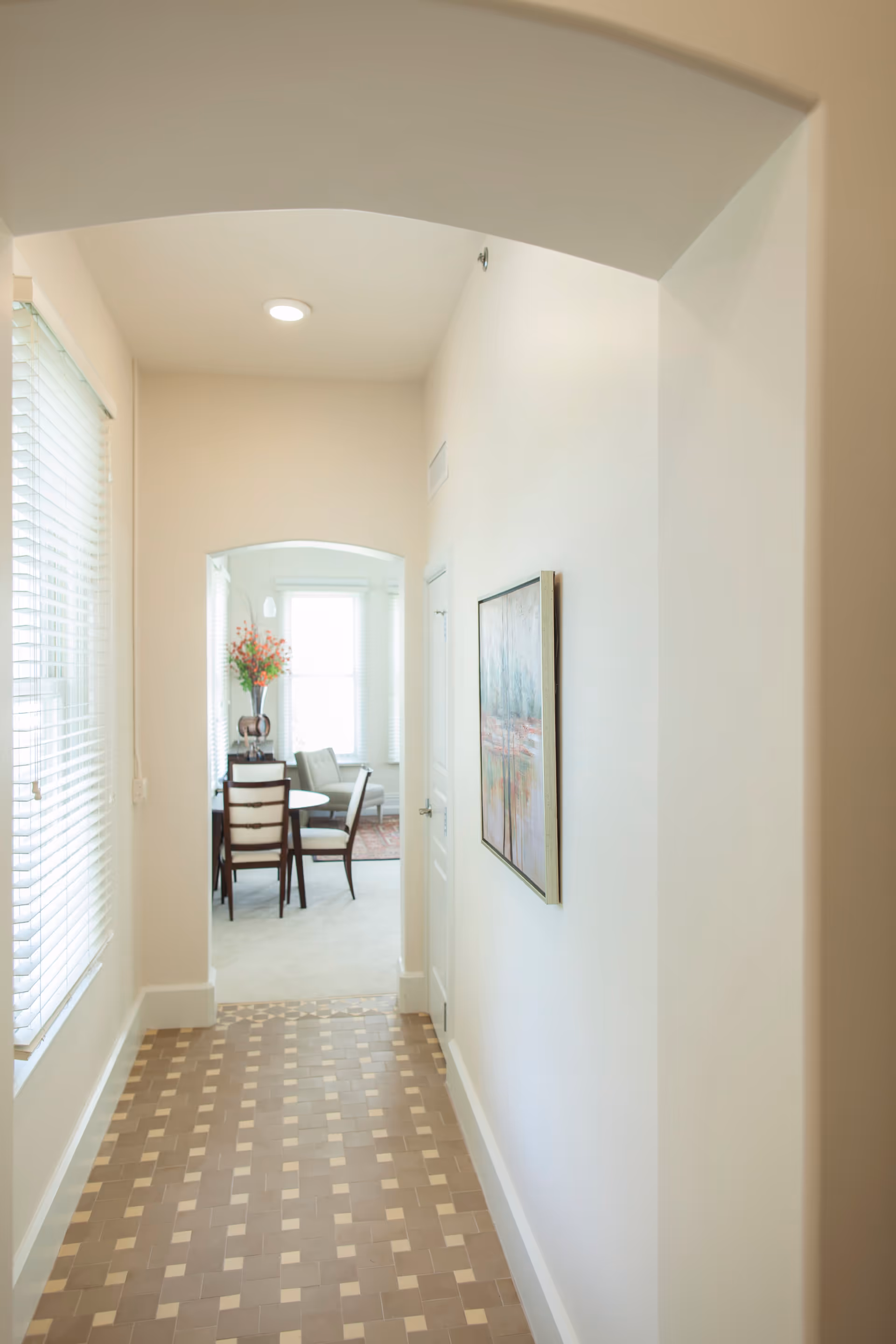 A bright hallway with patterned tile flooring and white walls leading to a dining area with a round table, chairs, and a vase with orange flowers. A framed painting hangs on the right wall, and large windows with blinds are on the left.