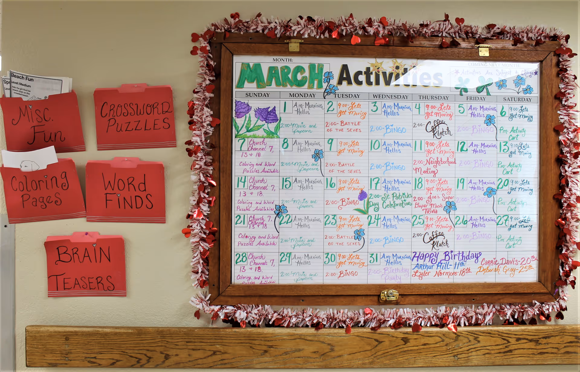 A bulletin board decorated with red and white garland displaying a March activities calendar with various events written in colorful markers. To the left of the board, red folders labeled Misc. Fun, Crossword Puzzles, Coloring Pages, Word Finds, and Brain Teasers are attached to the wall.