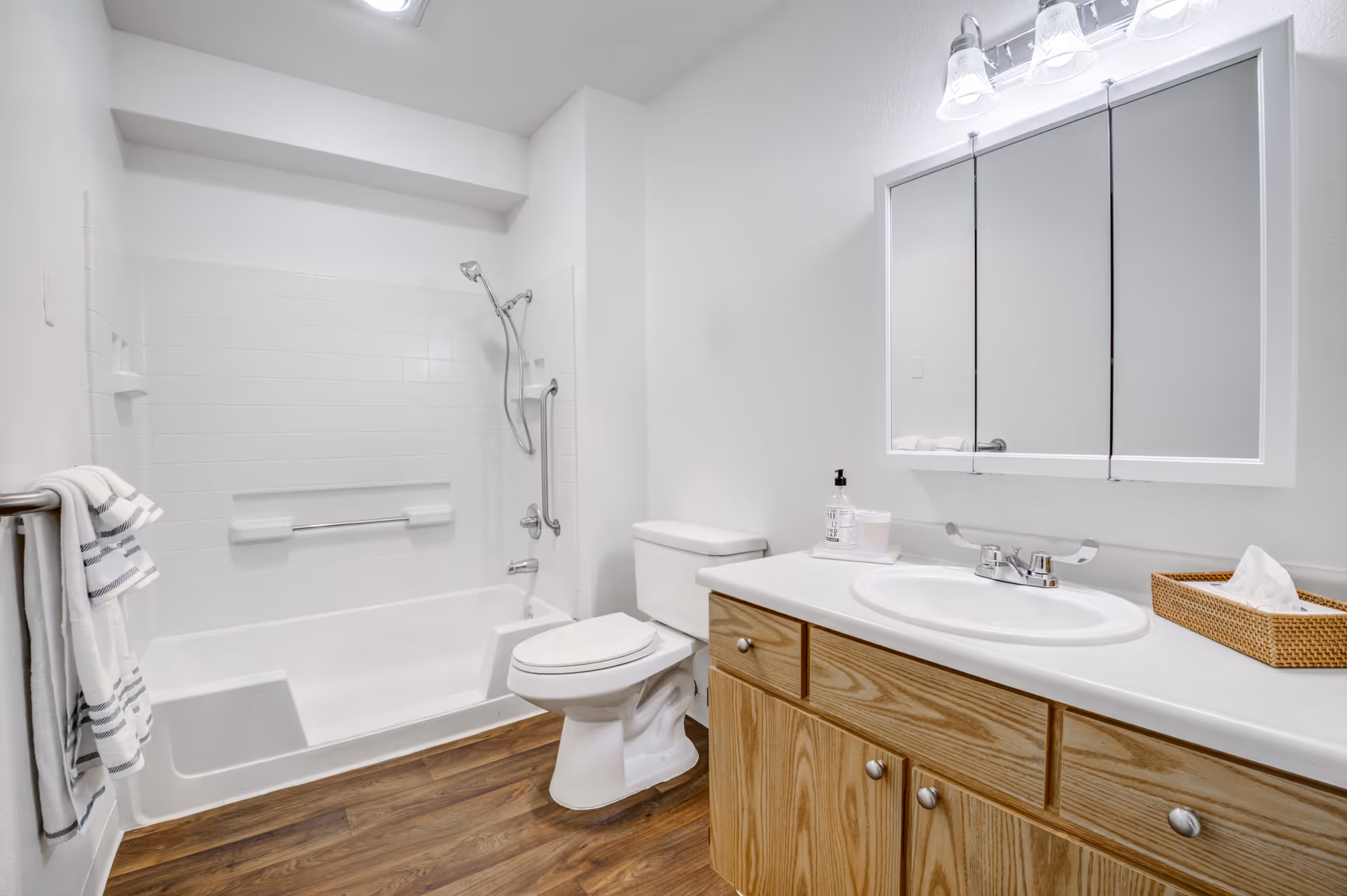 A clean and bright bathroom featuring a white bathtub with a handheld showerhead and grab bars, a white toilet, a wooden vanity with a white countertop and sink, a wall-mounted mirror cabinet with three sections, and a towel rack with white and gray striped towels. The floor has a wood-like finish.