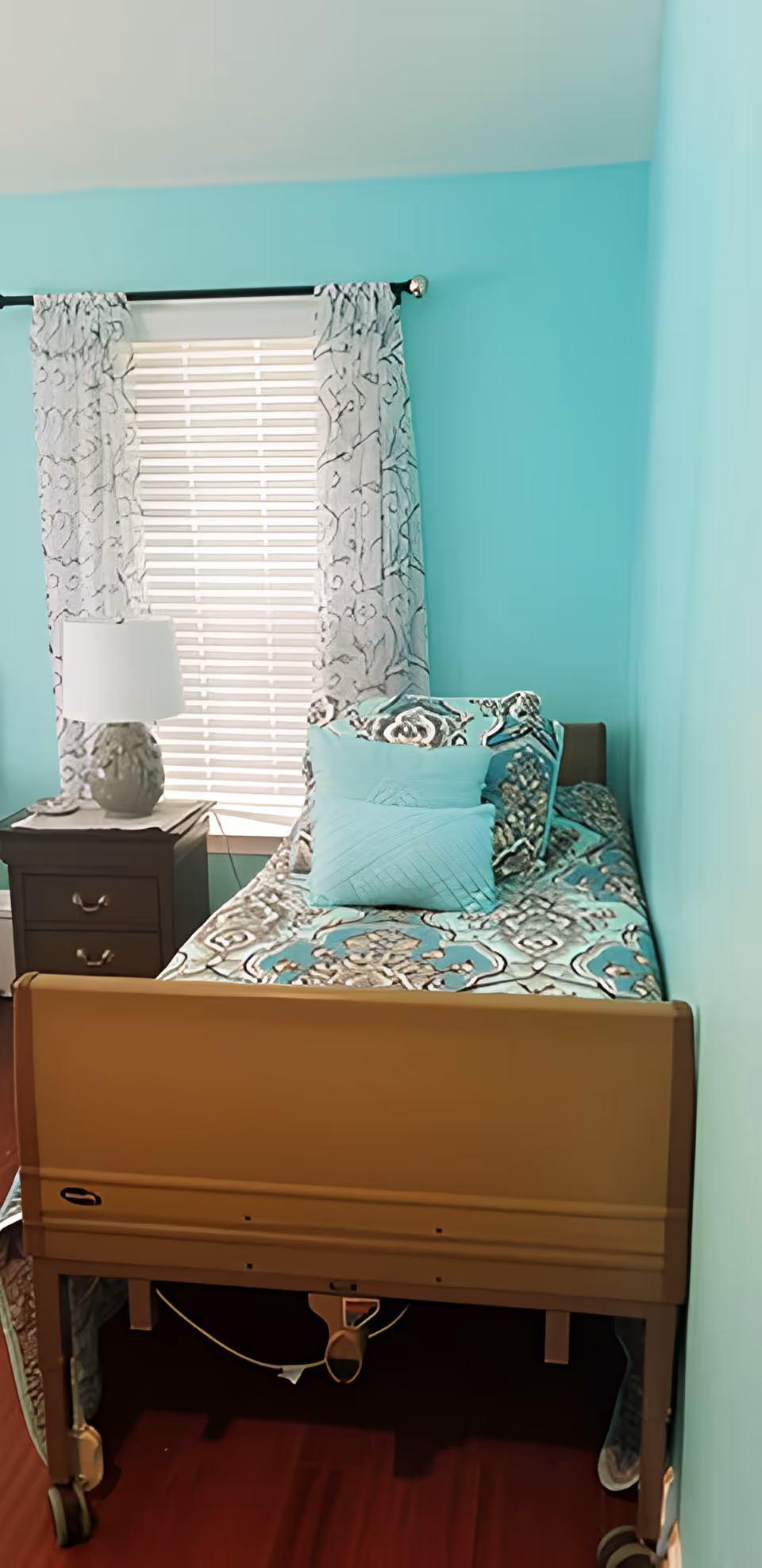 Single-occupancy bedroom with turquoise walls, an adjustable bed with patterned bedding, and a nightstand with a lamp by a window with blinds and curtains.