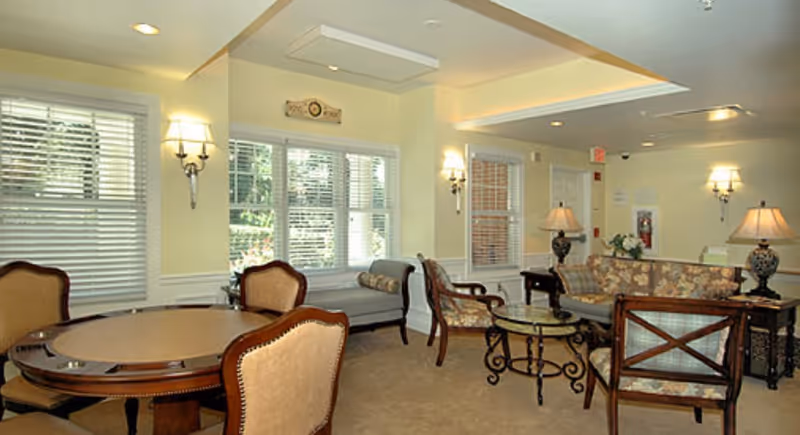 A cozy living room area with beige walls and carpet, featuring a round wooden table with four upholstered chairs, a gray cushioned bench under windows with white blinds, and a seating area with floral patterned sofas and armchairs around a glass-top coffee table. The room is softly lit by wall sconces and table lamps.