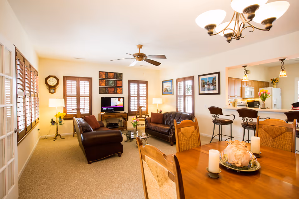 A bright and cozy living room with two brown leather sofas, a glass coffee table with flowers, a TV on a wooden stand, and several framed pictures on the walls. The room has multiple windows with wooden shutters, a ceiling fan, and lamps providing warm lighting. In the foreground, there is a wooden dining table with chairs, candles, and a decorative centerpiece. The kitchen is visible in the background with bar stools at a counter and pendant lights hanging above.