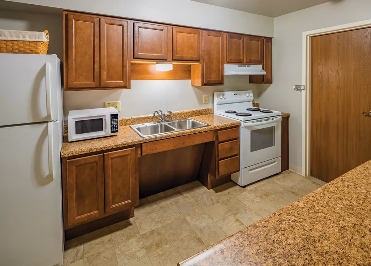 A kitchen with wooden cabinets, a white refrigerator, a microwave on the countertop, a double sink, and a white electric stove with an overhead vent. The floor is tiled and there is a wooden door on the right side.